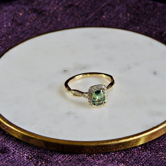 Gold ring with a cushion-cut green gemstone center and diamond halo, displayed on a round white marble dish with a gold rim against a purple fabric background.