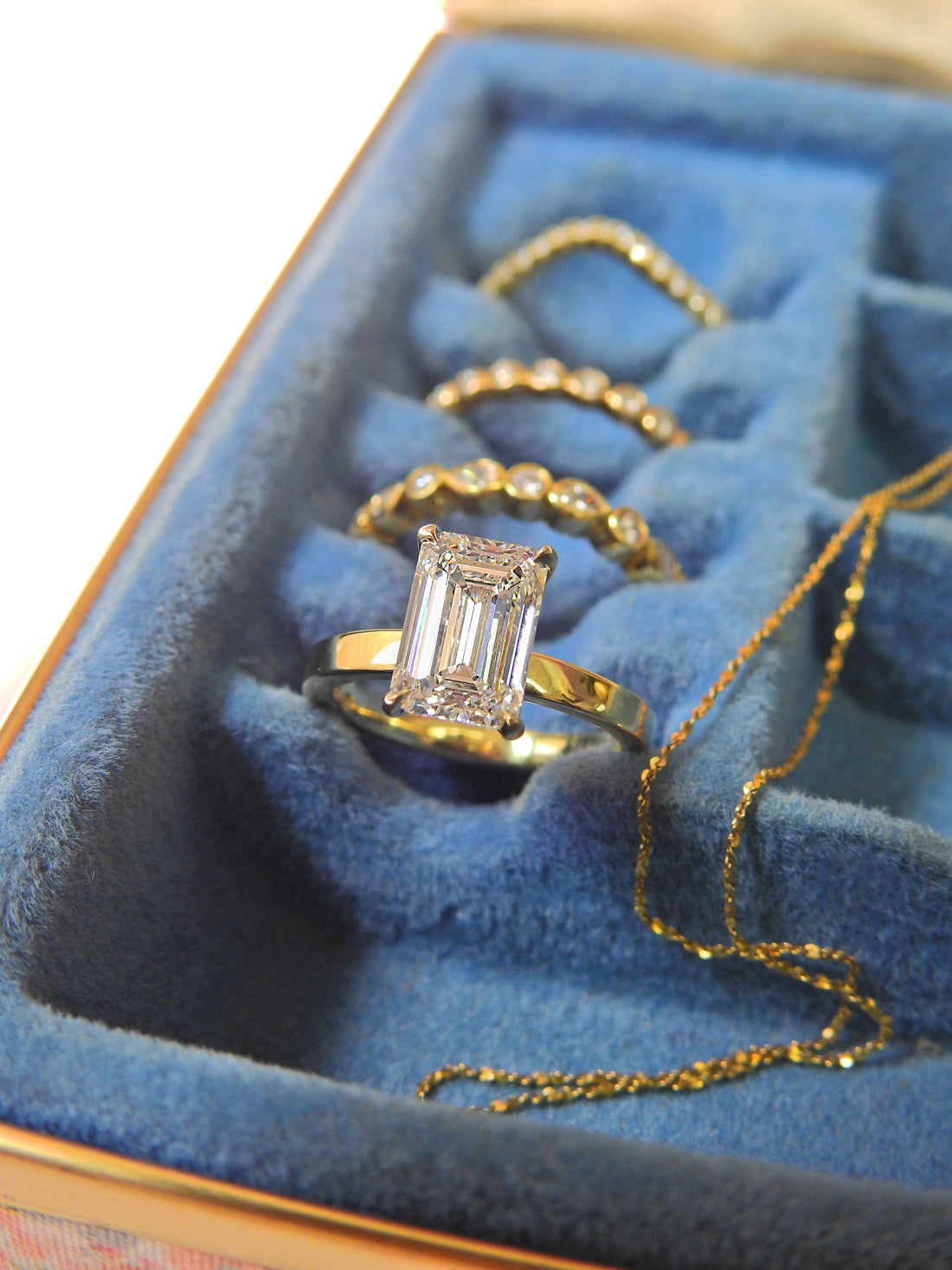 Emerald-cut diamond engagement ring in a yellow gold band by Ethan Lord Jeweler, displayed in a blue velvet jewelry box with gold rings and chain.