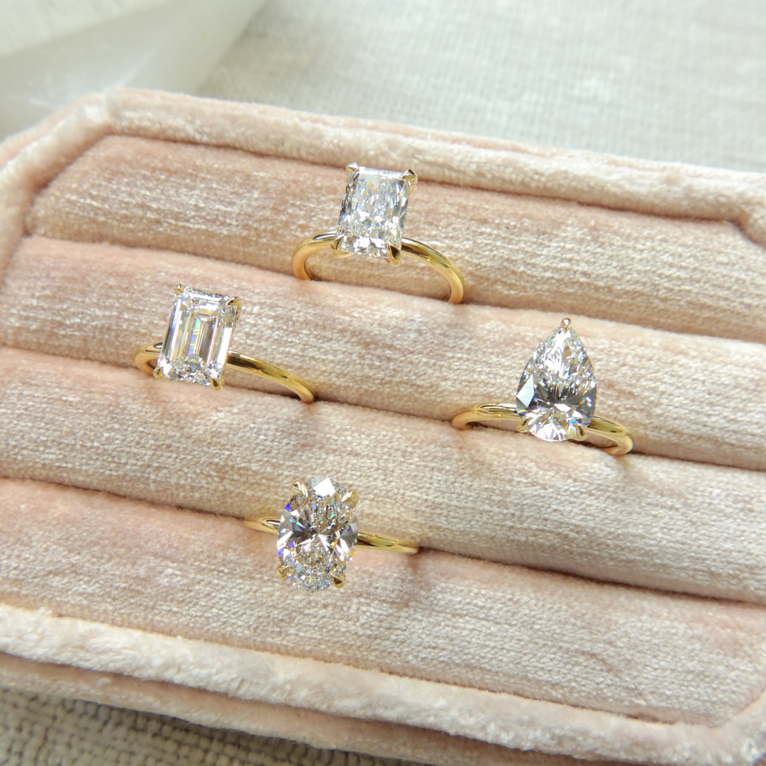 Four solitaire diamond engagement rings featuring emerald, radiant, pear, and oval cuts, set in yellow gold bands and displayed in a beige velvet jewelry box at Ethan Lord Jewelers.