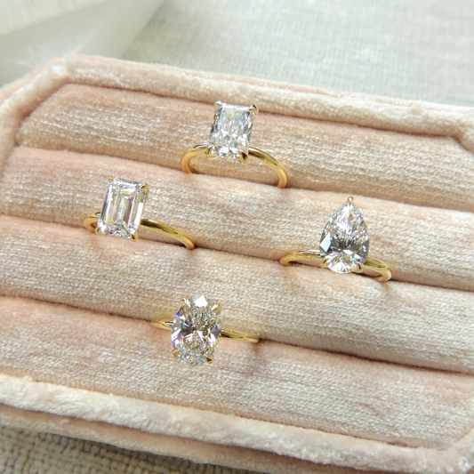 Four solitaire diamond engagement rings featuring emerald, radiant, pear, and oval cuts, set in yellow gold bands and displayed in a beige velvet jewelry box at Ethan Lord Jewelers.