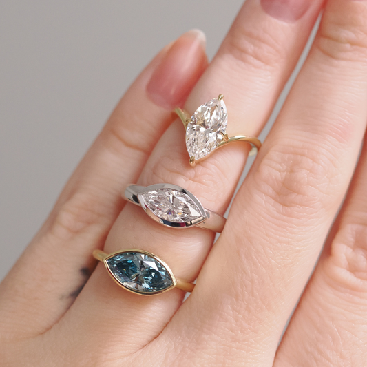 Three marquise-cut gemstone rings worn on a hand, featuring a clear diamond solitaire in yellow gold, a bezel-set clear diamond in white metal, and a bezel-set deep blue gemstone in yellow gold, showcasing custom ring designs at Ethan Lord Jewelers.