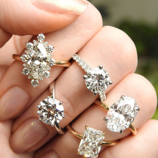 Close-up of a hand wearing five diamond engagement rings in different styles — marquise halo, round solitaire, oval cut, emerald cut, and round with pavé band.