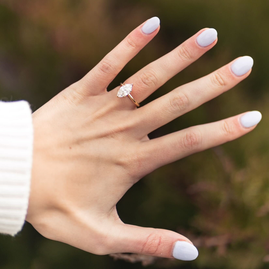 Hand wearing a gold engagement ring with a marquise-cut diamond against a blurred outdoor background.