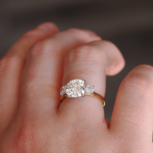 Close-up of a hand wearing a three-stone engagement ring featuring a cushion-cut diamond center with two square-cut side stones set on a yellow-gold band.