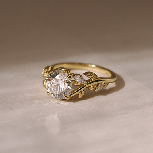 Gold engagement ring with a round-cut diamond center stone, accented by delicate leaf/vine detailing and small marquise-shaped diamonds on a soft neutral background.