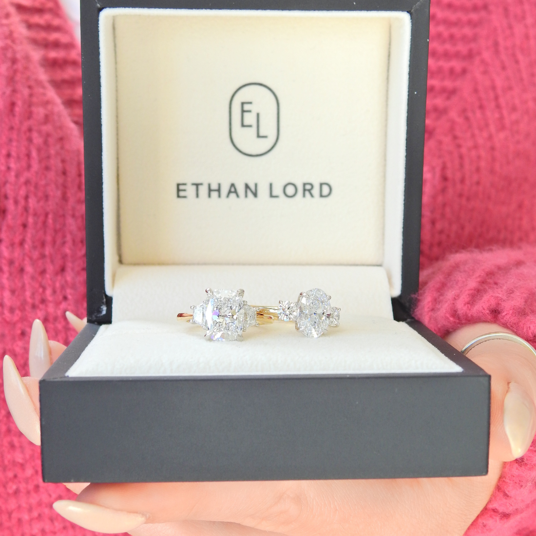 Open Ethan Lord ring box held in hand, displaying two diamond engagement rings with sparkling center stones on a white cushion.