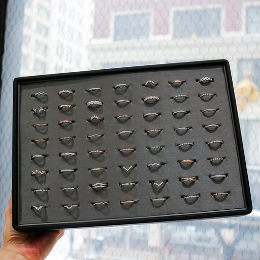 A hand holds a black display tray filled with dozens of delicate diamond bands and stacking rings, arranged in neat rows against a softly blurred window backdrop.