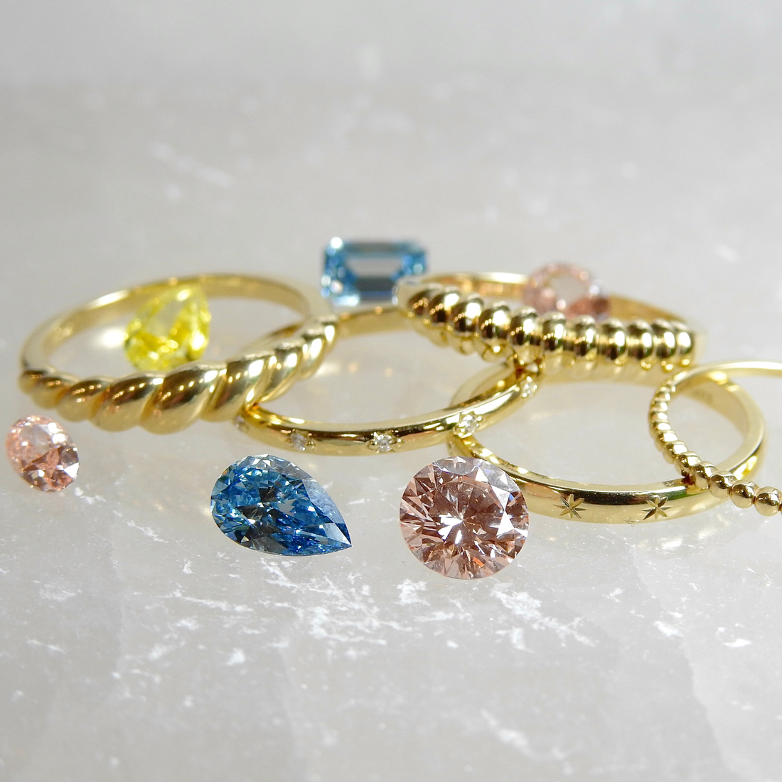 Gold rings in assorted styles displayed on a stone surface with loose blue, pink, and yellow gemstones in front.