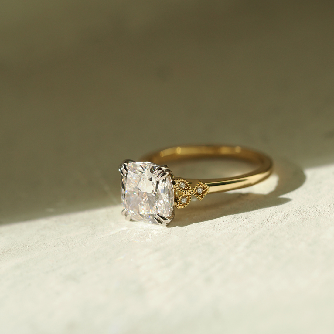 Vintage-inspired gold engagement ring with a cushion-cut diamond and delicate side detailing on a soft neutral background.